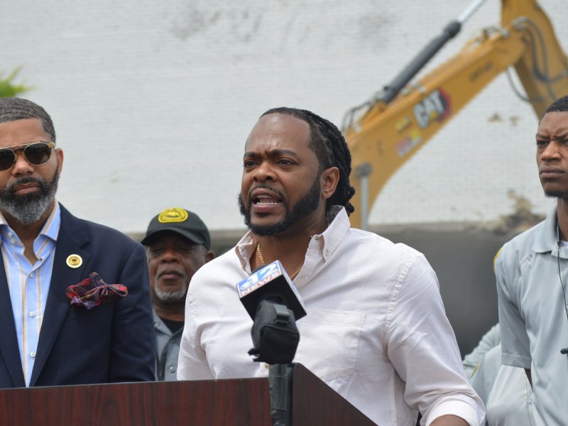 Three men stand at a podium with a mic and behind them is a large yellow demolition truck
