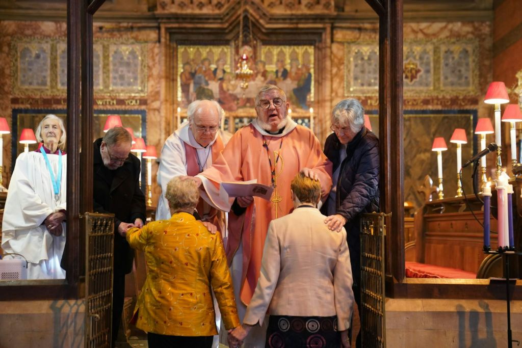 Two women in formal jackets kneel before a clergyman in an orange robe