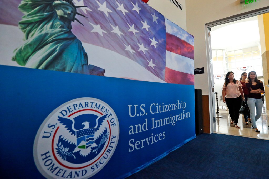 Woman walking out of a room with a backdrop that says U.S. Citizenship and Immigration Services right next to them