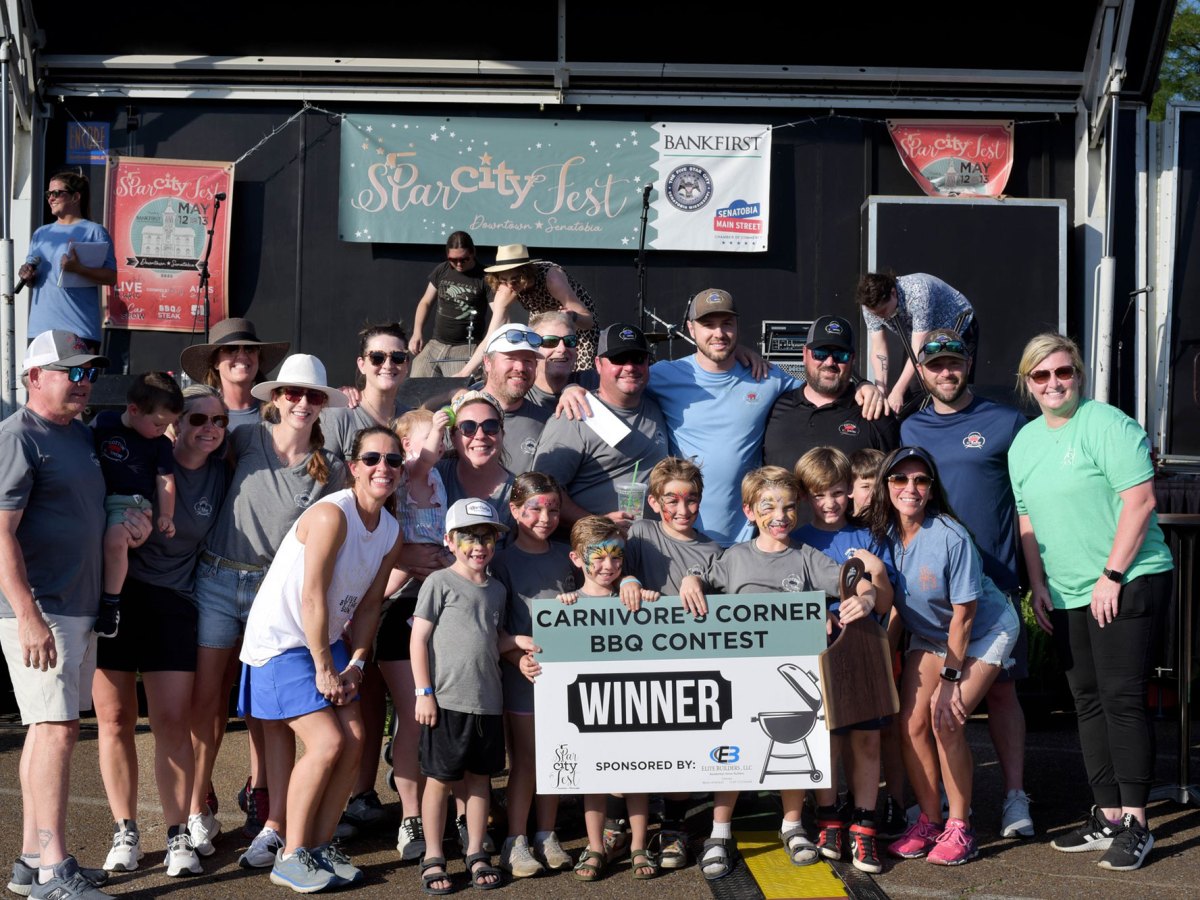 Many people gathered in front of an outdoor stage holding a sign that reads Carnivore's Corner BBQ Contest Winner