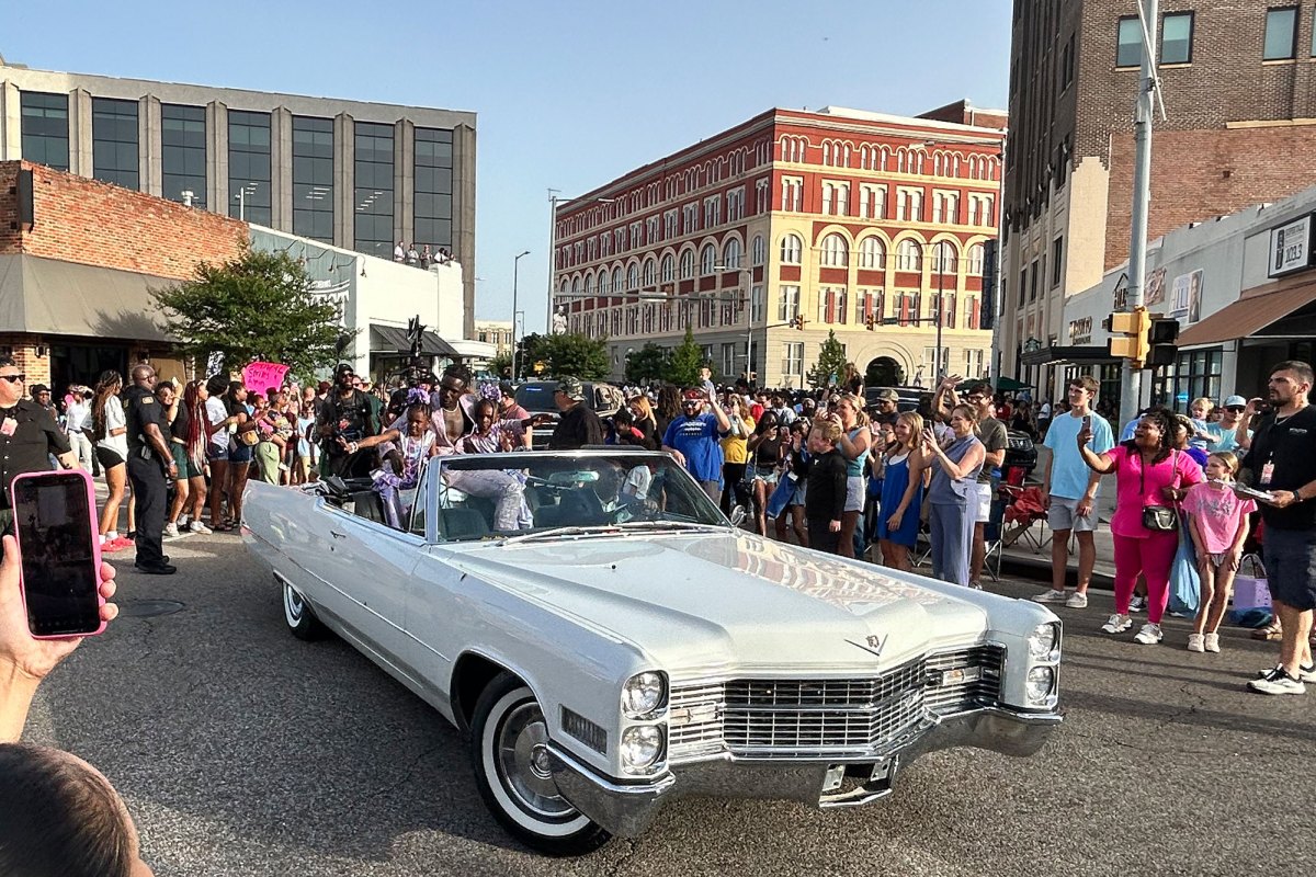 A man sits in the back of a white convertible car with two small children as they drive through a parade of onlookers