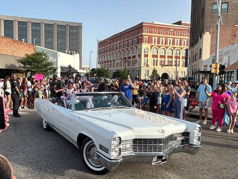 A man sits in the back of a white convertible car with two small children as they drive through a parade of onlookers