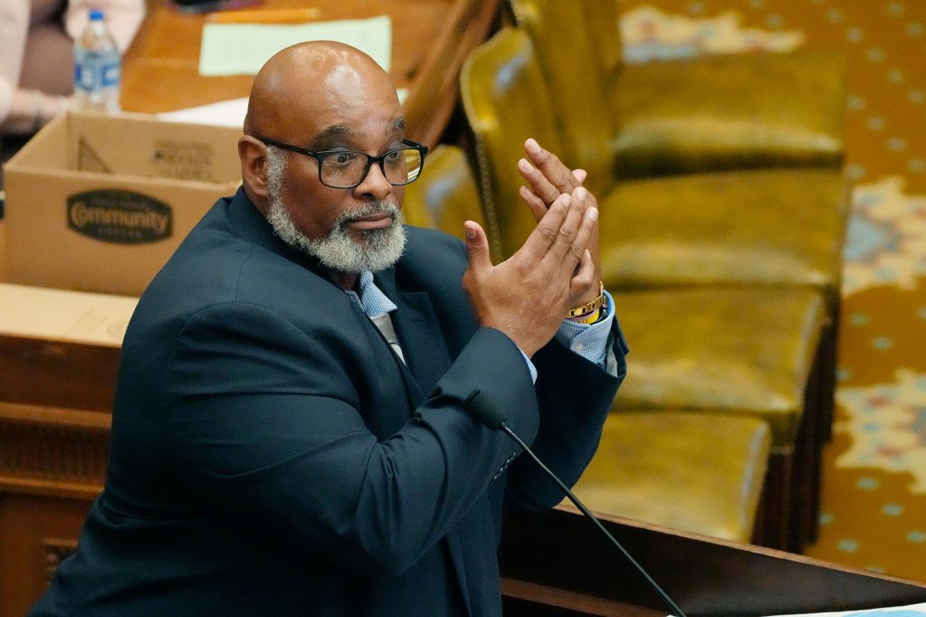 A man gestures at his hands while speaking at a podium