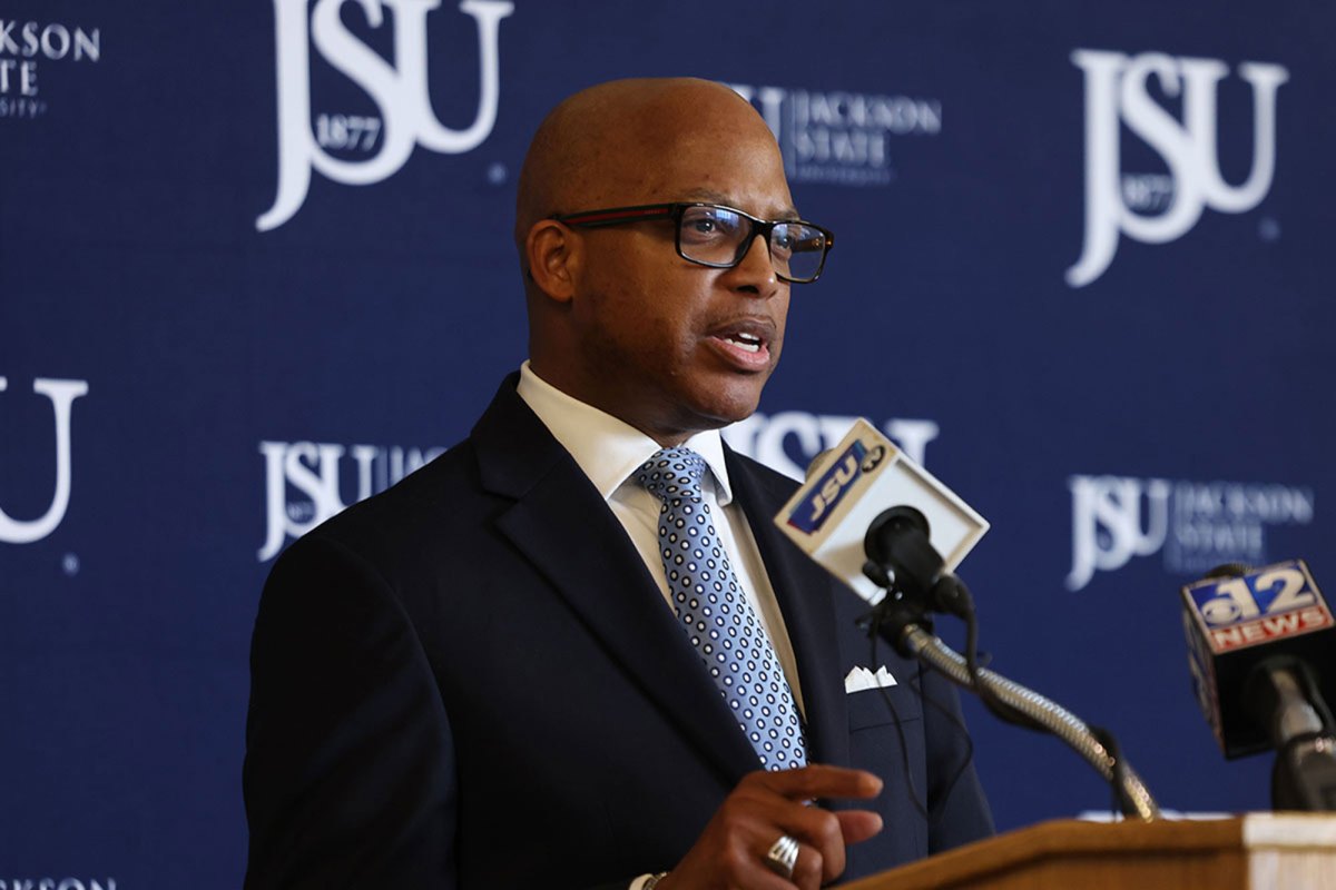 A man wearing a black jacket with a blue tie with white dots speaks at a podium in front of a blue backdrop with JSU written on it