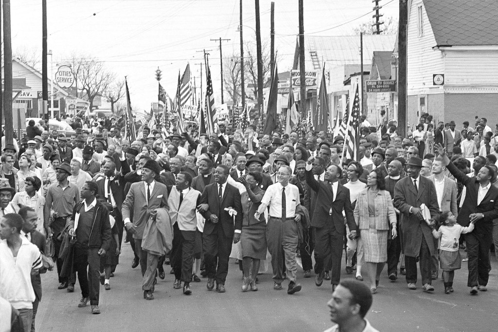 Dr. Martin Luther King marches with a large crown carrying U.S. Flags down a main road