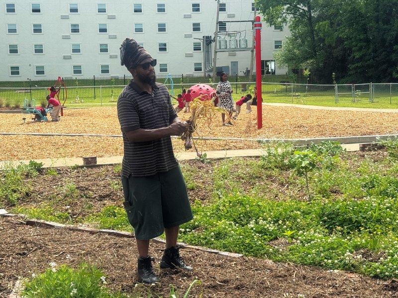 A man in black standing in a garden and behind him a playground with children wearing red