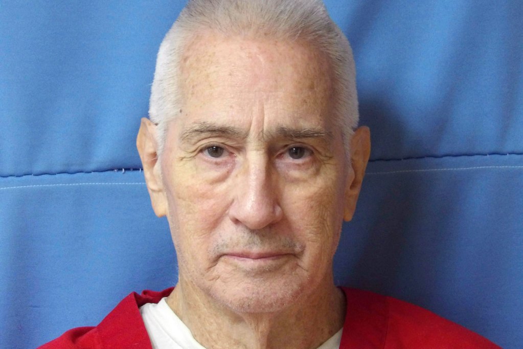 An older man in a red prison jumpsuit against a blue wall