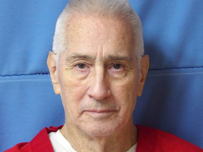 An older man in a red prison jumpsuit against a blue wall