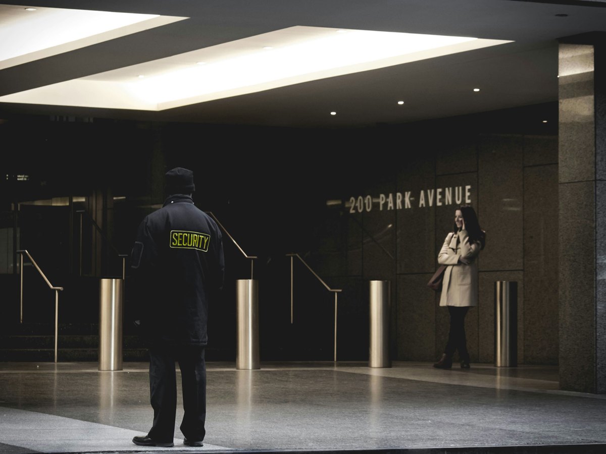 Security guard standing in a building in all black with a woman standing off to his right in a tan jacket