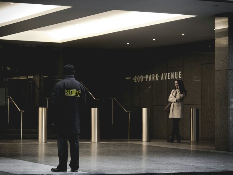Security guard standing in a building in all black with a woman standing off to his right in a tan jacket