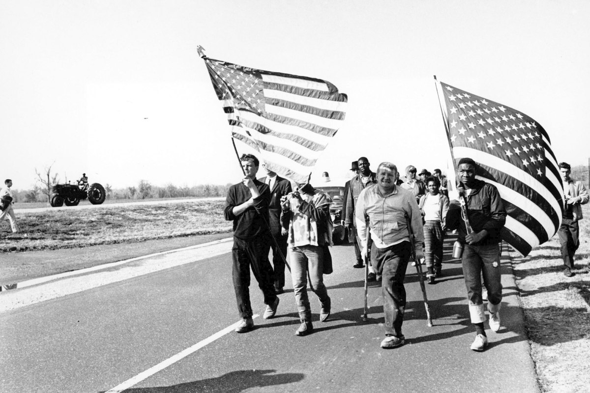 A black and white photo of people marching down a road, carrying U.S. flags