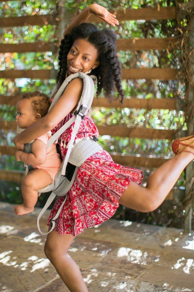 A woman in a red dress with a baby strapped to her does a dance move while smiling