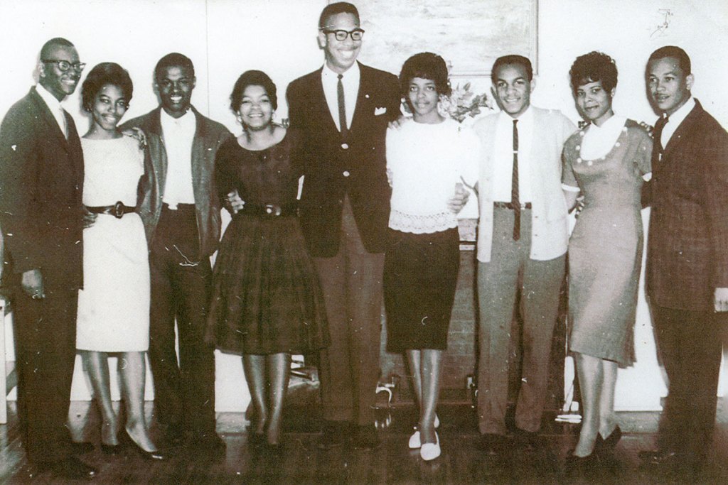 an old 1960s era black and white photo shows five Black men and four Black women standing together posing for a photo