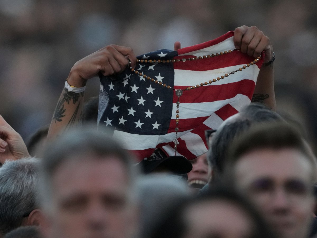 Closeup of hands holding up a US flag and a catholic rosary above the crowd