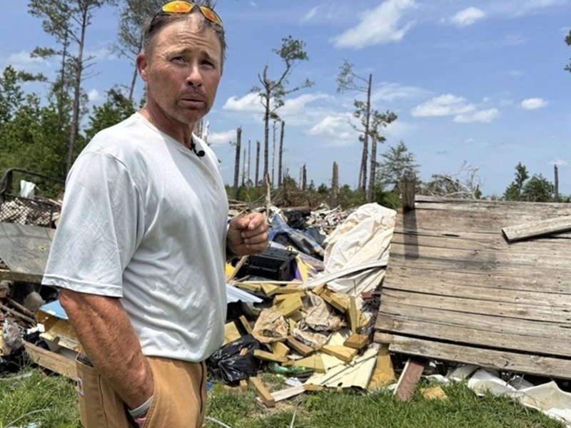 Man in grey shirt and khaki's stand in front of a pile of debris in an open field