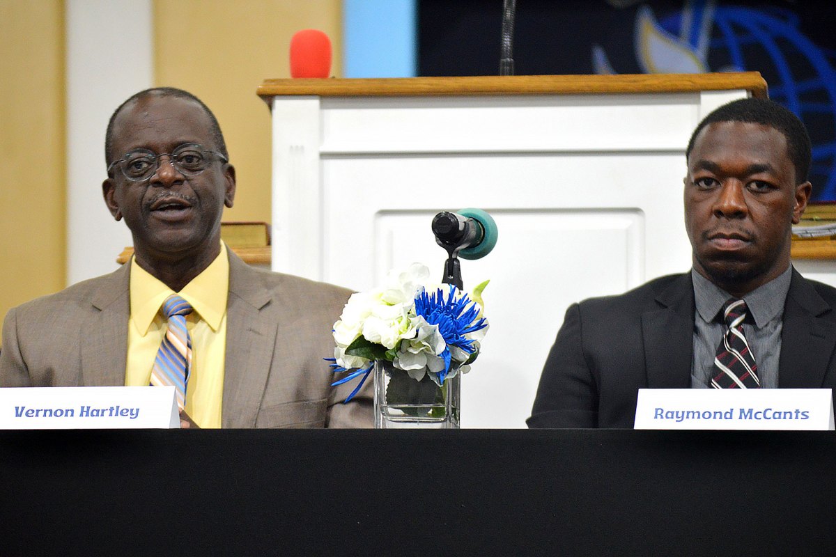 Two seated men in suits behind signs that label them as Vernon Hartley on the left and Raymond McCants on the right
