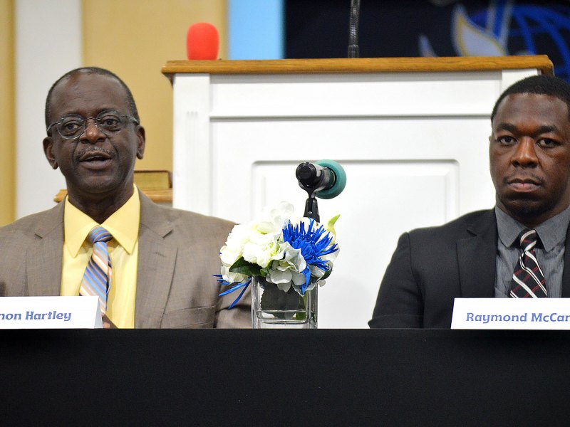 Two seated men in suits behind signs that label them as Vernon Hartley on the left and Raymond McCants on the right
