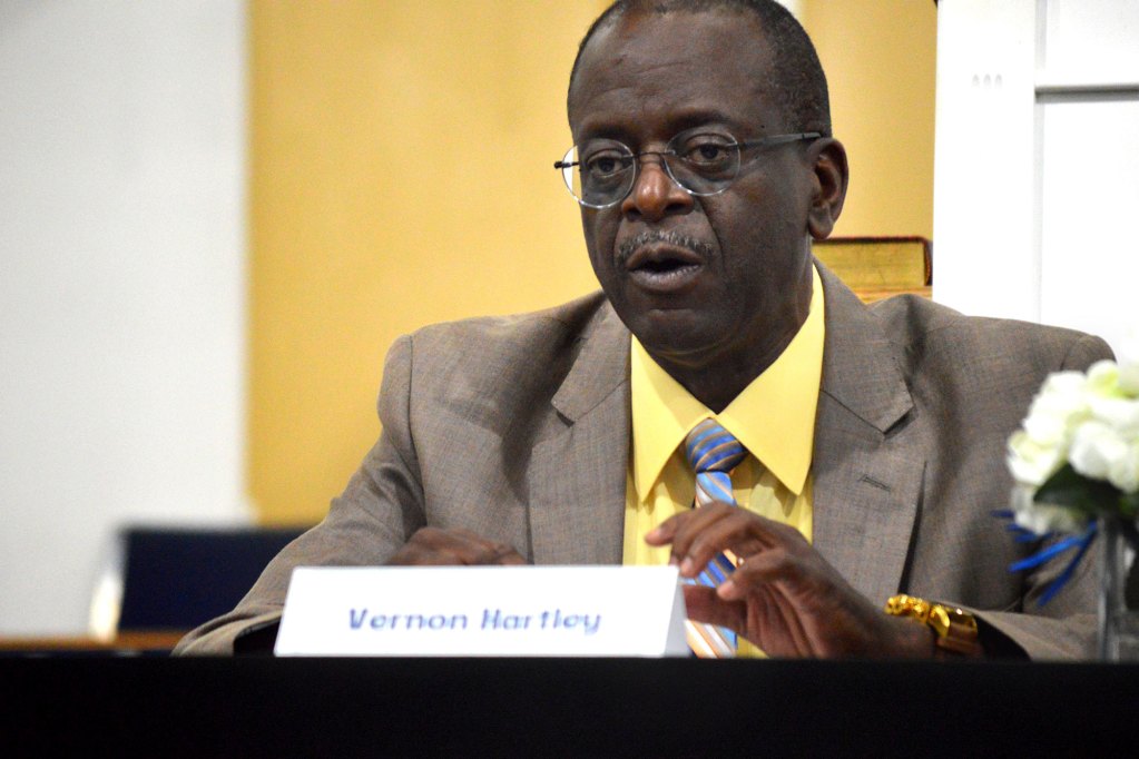 A man speaking behind a sign that labels him as Vernon Hartley