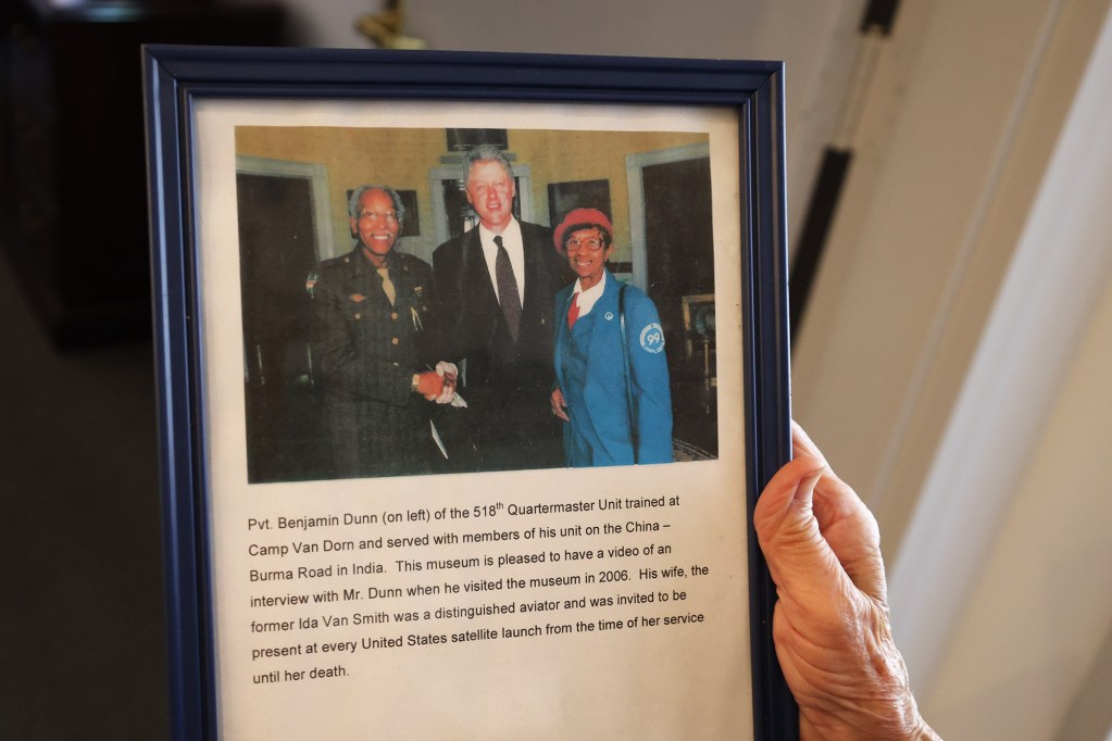 A WW2 museum display featuring President Bill Clinton with two people in uniform. The text below reads: Pvt. Benjamin Dunn (on left) of the 51gth Quartermaster Unit trained at Camp Van Dorn and served with members of his unit on the China - Burma Road in India. This museum is pleased to have a video of an interview with Mr. Dunn when he visited the museum in 2006. His wife, the former Ida Van Smith was a distinguished aviator and was invited to be present at every United States satellite launch from the time of her service until her death.