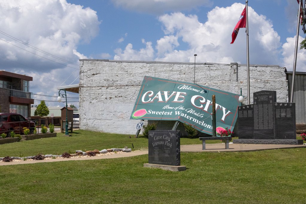A green sign that says welcome to cave city sits in front of a white brick building
