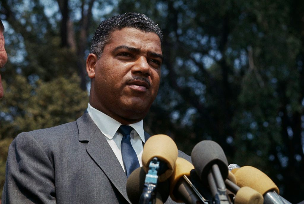Black man wearing suit stands before microphones