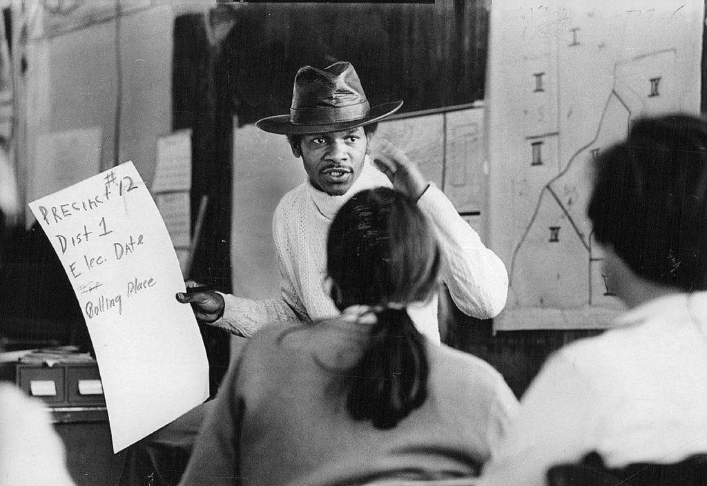 Young Black man wearing a cowboy hat speaks to a group while holding a poster with voting information