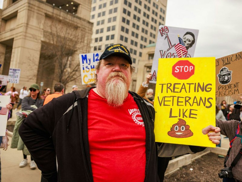 A man standing at a protest with a yellow sign that says "Stop Treating Veterans Like Poop"