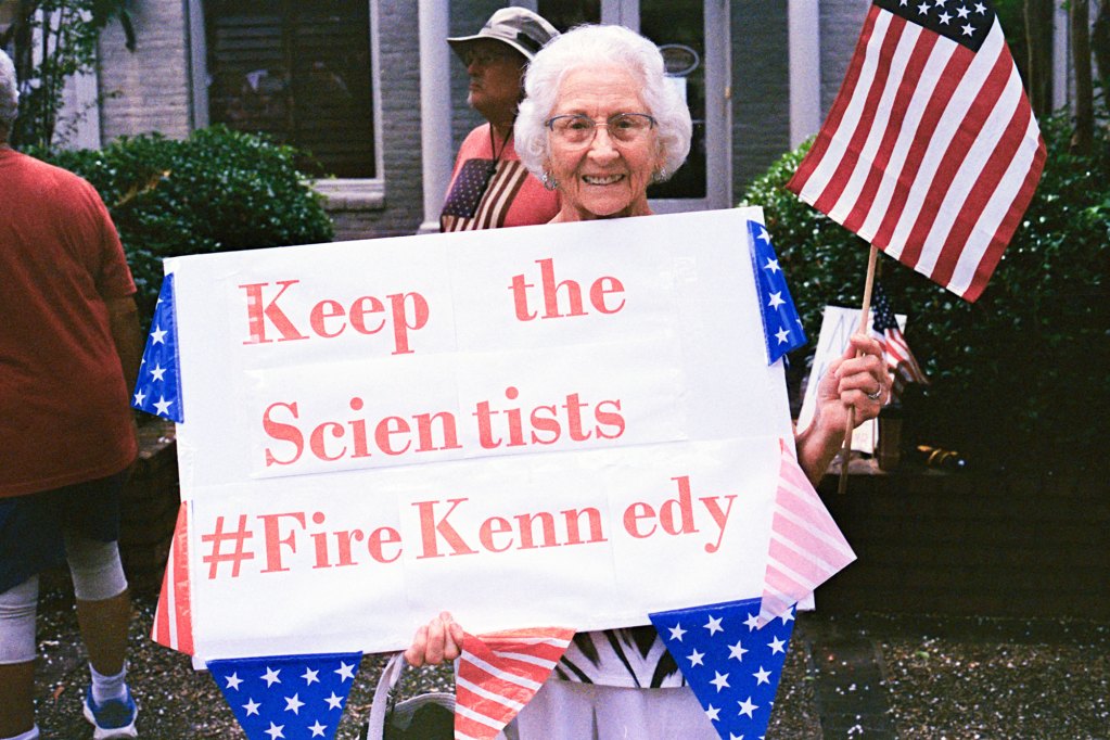 An older woman holds a US flag and a sign that reads 'Keep the Scientists #FireKennedy"