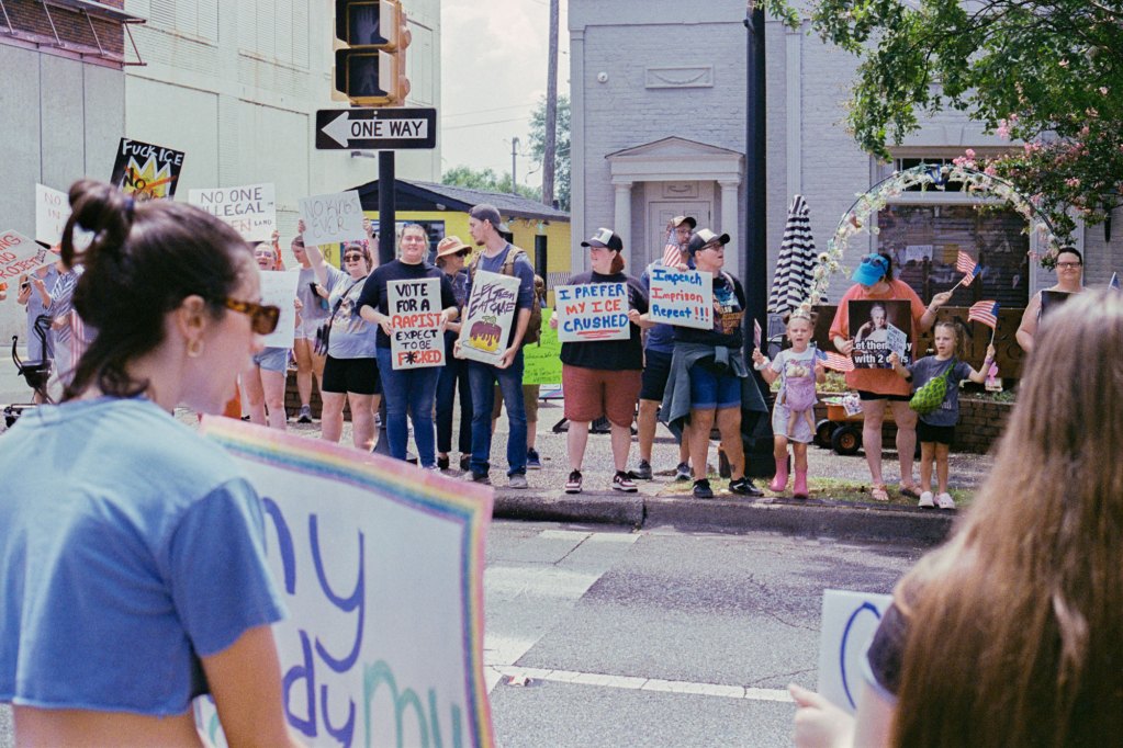 Protesters stand on both sides of the street holding signs, some read "Impeach Imprison repent" and "I prefer my ice crushed"