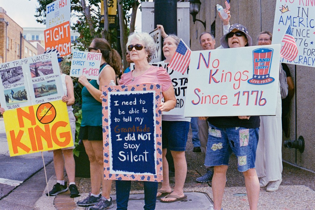 Protesters hold signs that read "No kings since 1776" and "I need to be able to tell my grandkids I did not stay silent"