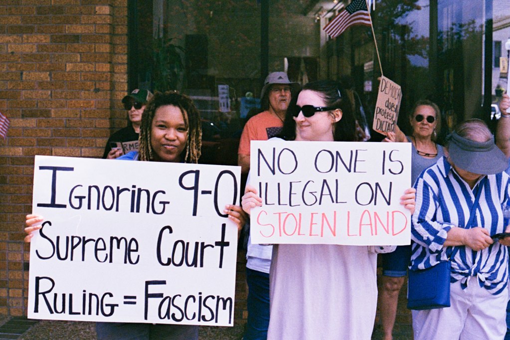 Protesters hold signs that read "Ignoring 9-0 Supreme Court Rulling = Facism" and "No one is illegal on stolen land"