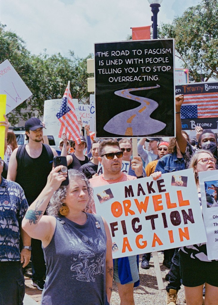 Protesters hold signs that read "The road it fascism is lined with people telling you to stop overreacting" and "Make Orwell fiction again"