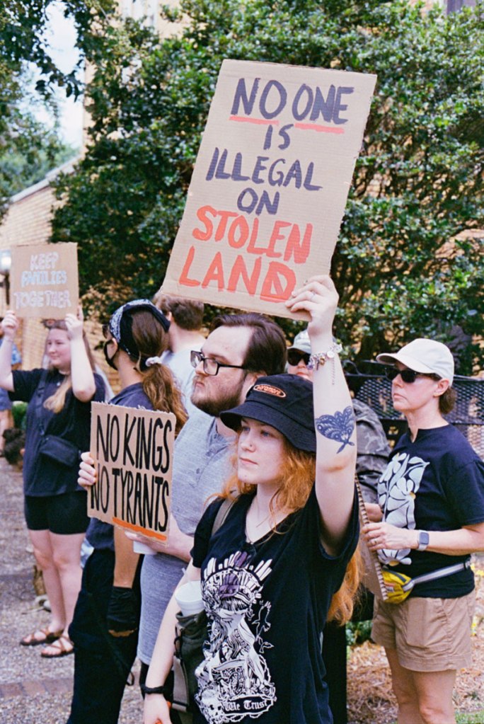 Protestors hold signs, the big one reads "No one is illegal on stolen land"