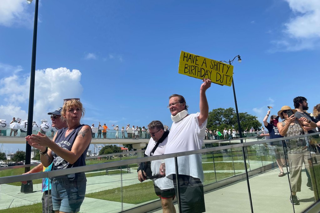 Protesters walk on a bridge holding signs, one reads "Have a shitty birthday DJT"