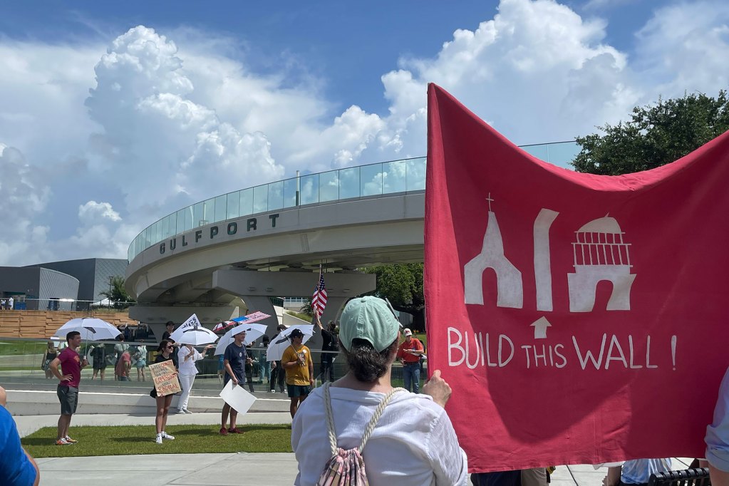 Protesters stand outside holding signs, one is large and red and says Build This Wall