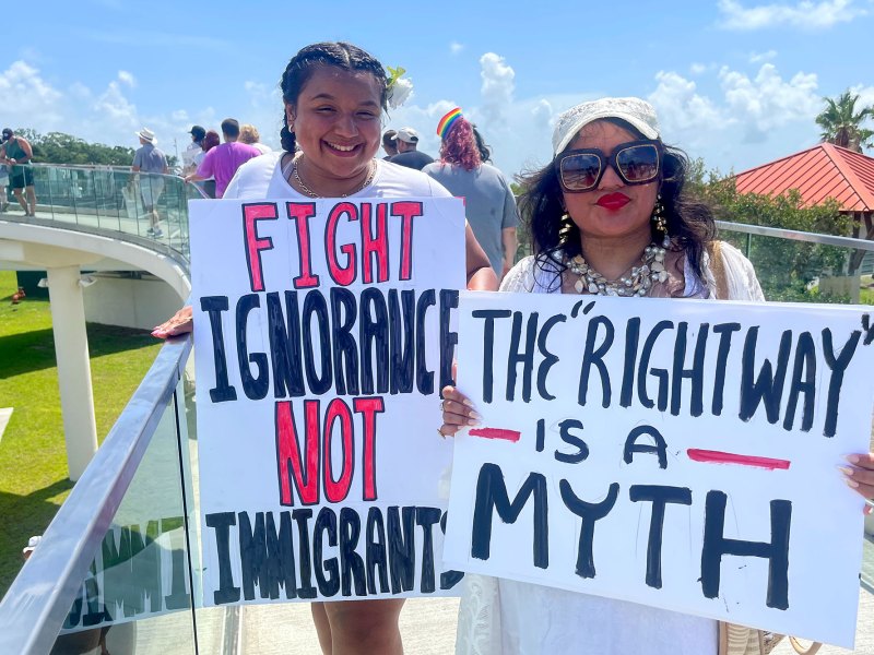 Two protesters hold signs on a bridge. They read "Fight Ignorance Not Immigrants" and "The 'right way' is a myth"
