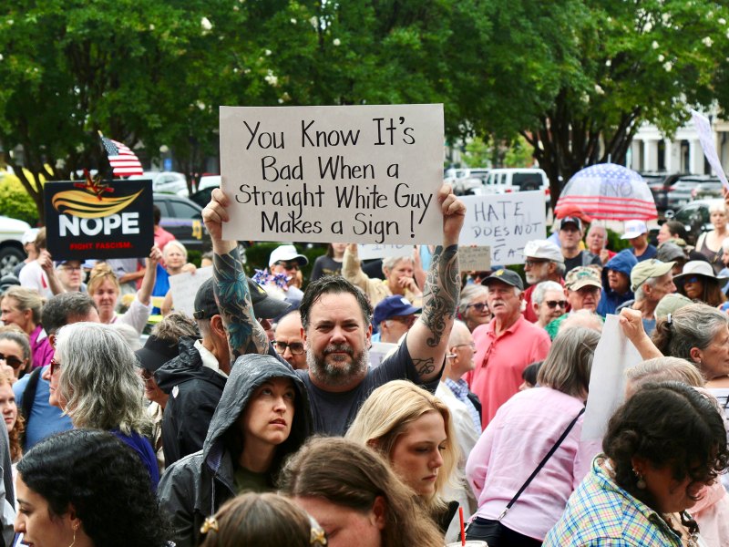 A crowd of protesters carry signs, one reads "You know it's bad when a straight white guy makes a sign!"