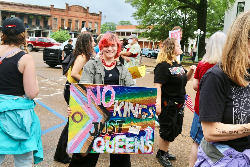 A crowd of protesters carry signs, one reads "No Kings just queens"