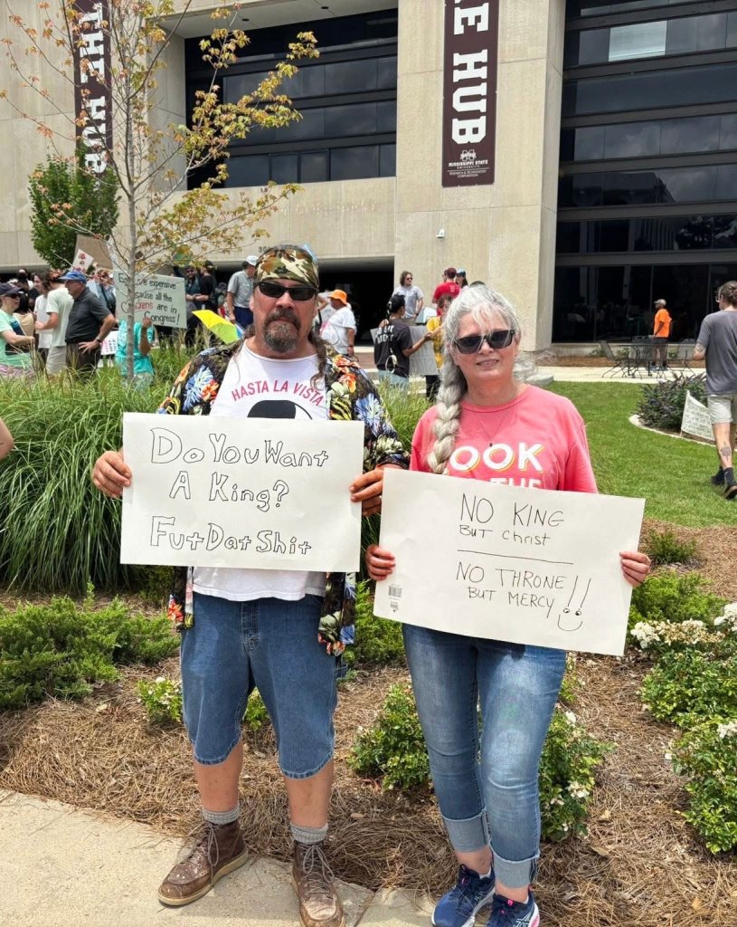 Protesters hold up signs, one reads "Do you want a king? Fut dat shit" and the other "No King but Christ - No throne but mercy!"