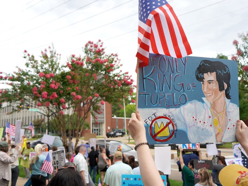 A crowd of protesters carry signs, one reads "Only king of Tupelo" with a painting of Elvis