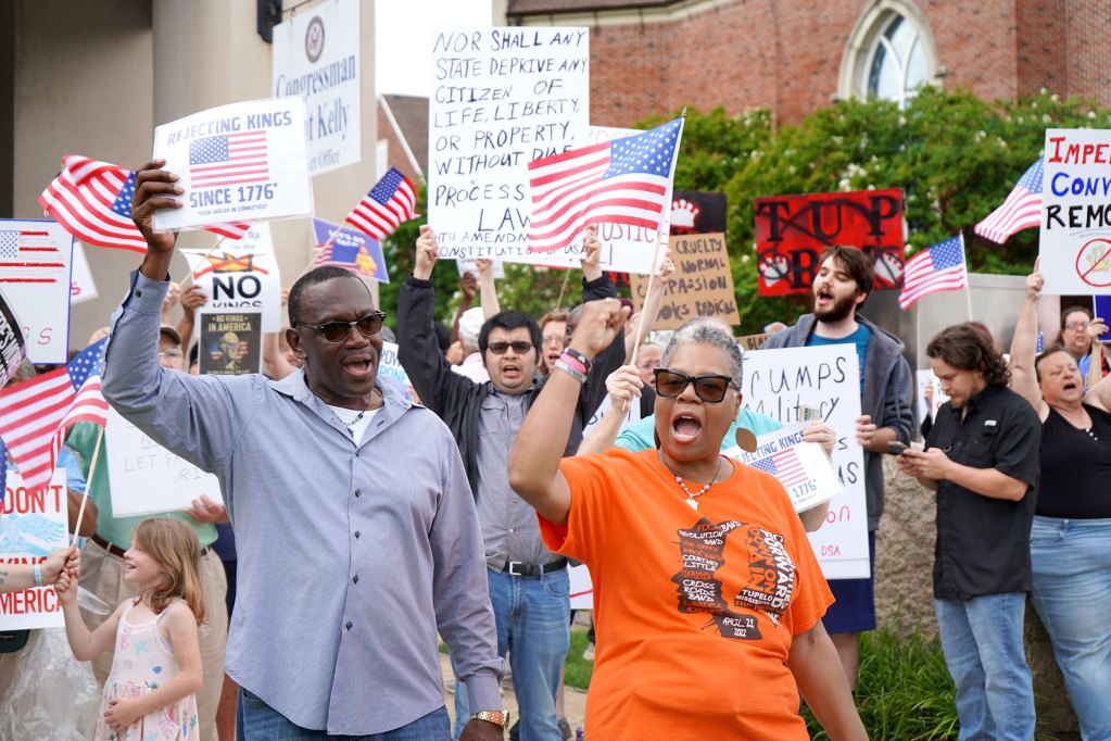 A crowd of protesters carry US flags and signs, one reads "Rejecting kings since 1776"