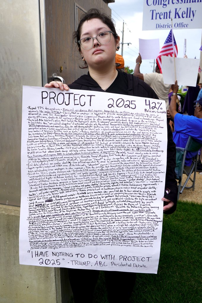 A crowd of protesters carry signs, one carrying a very long and text heavy sign about Project 2025