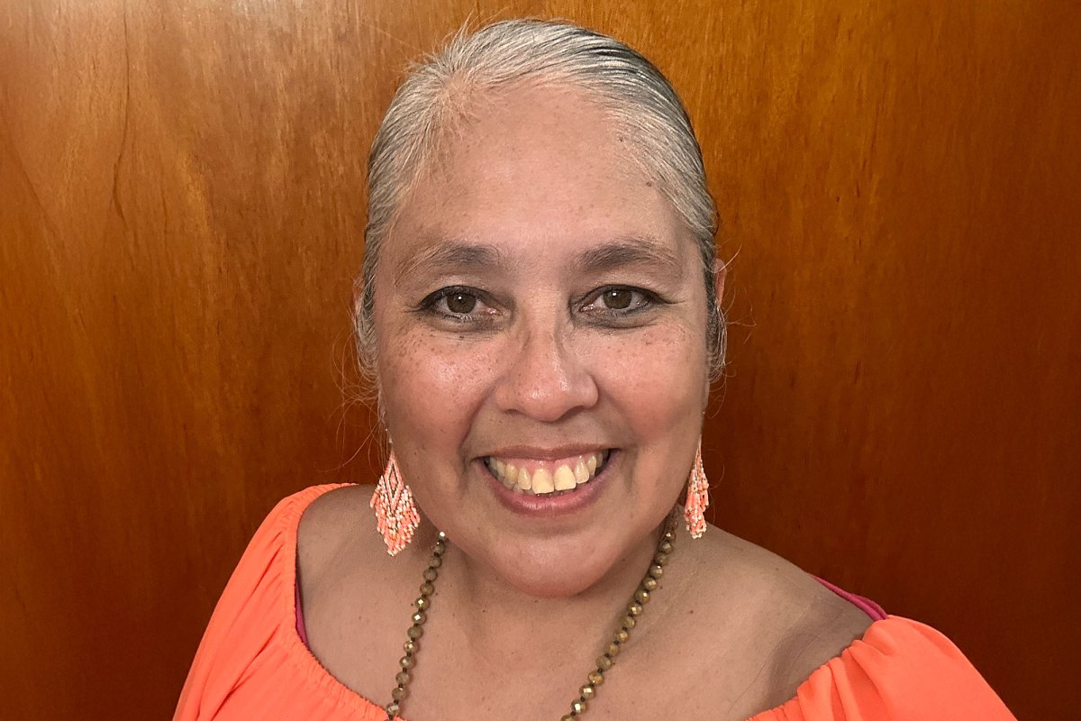 A woman in an orange top and matching orange beaded earrings smiles in front of a wooden wall