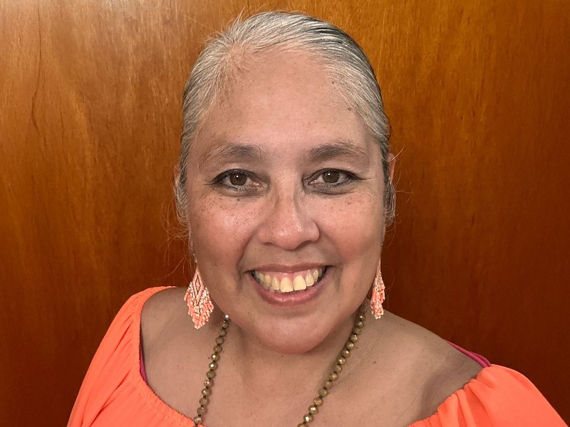 A woman in an orange top and matching orange beaded earrings smiles in front of a wooden wall