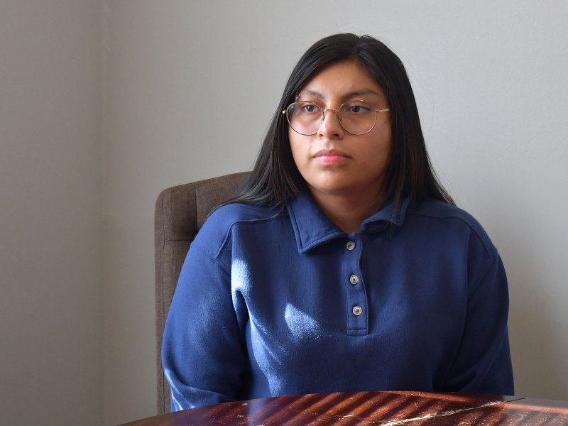 A young woman with long dark hair and glasses sits at a table near a window. She is wearing a blue collared shirt and looking slightly to the side.