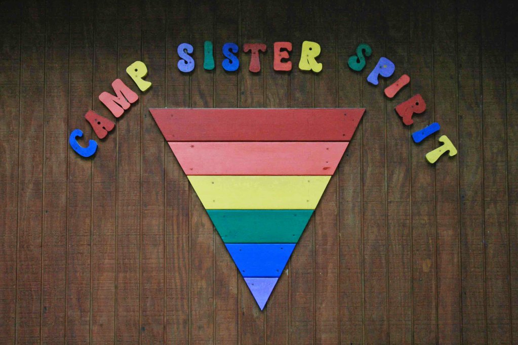 An upside down rainbow triangle on a wooden wall, above it in rainbow colors is the name Camp Sister Spirit
