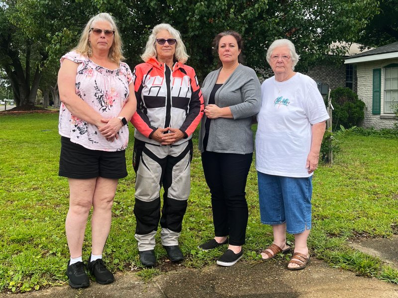 Four women stand side by side along the edge of a front yard driveway