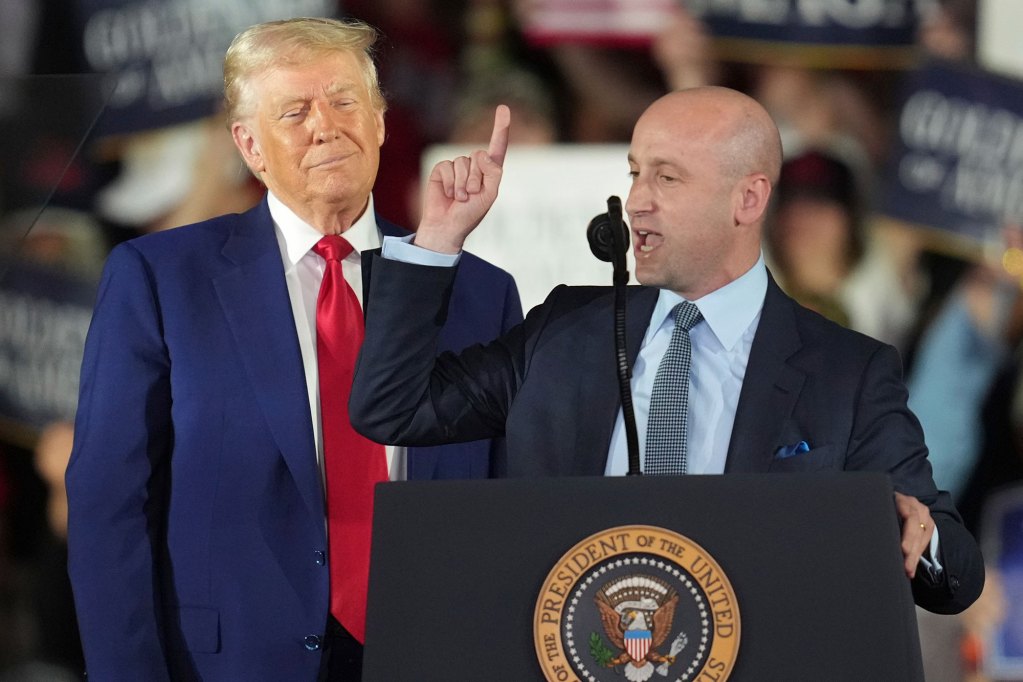 Donald Trump smiles on as Stephen Miller talks from the podium, finger pointing up