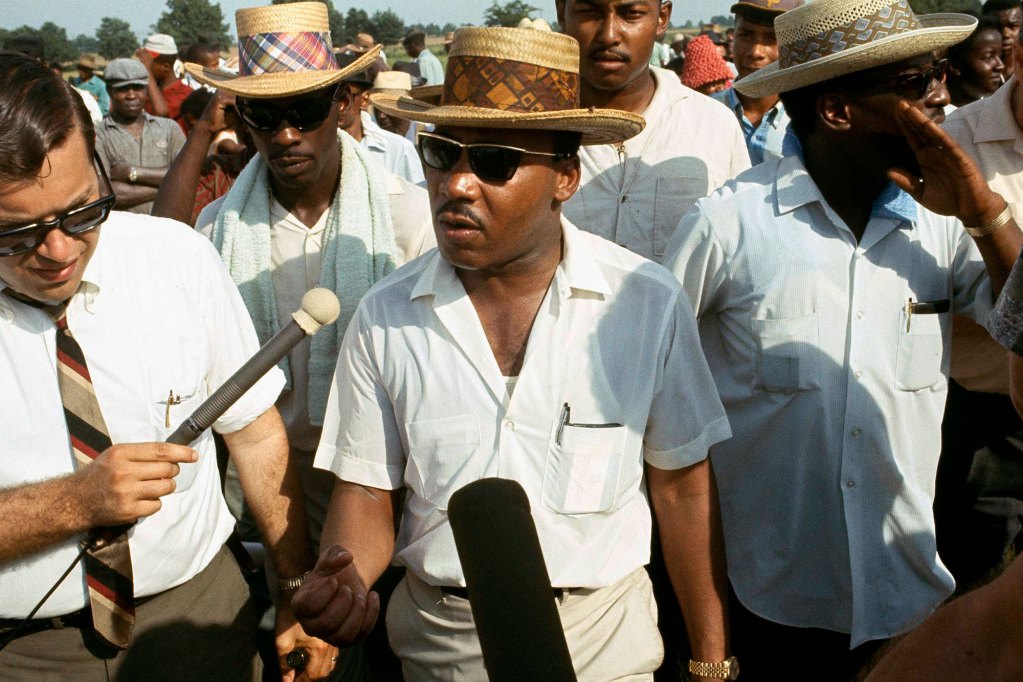 Martin Luther King Jr. marches wearing a straw hat and red shirt with a group of other men while a man holds a microphone up to him