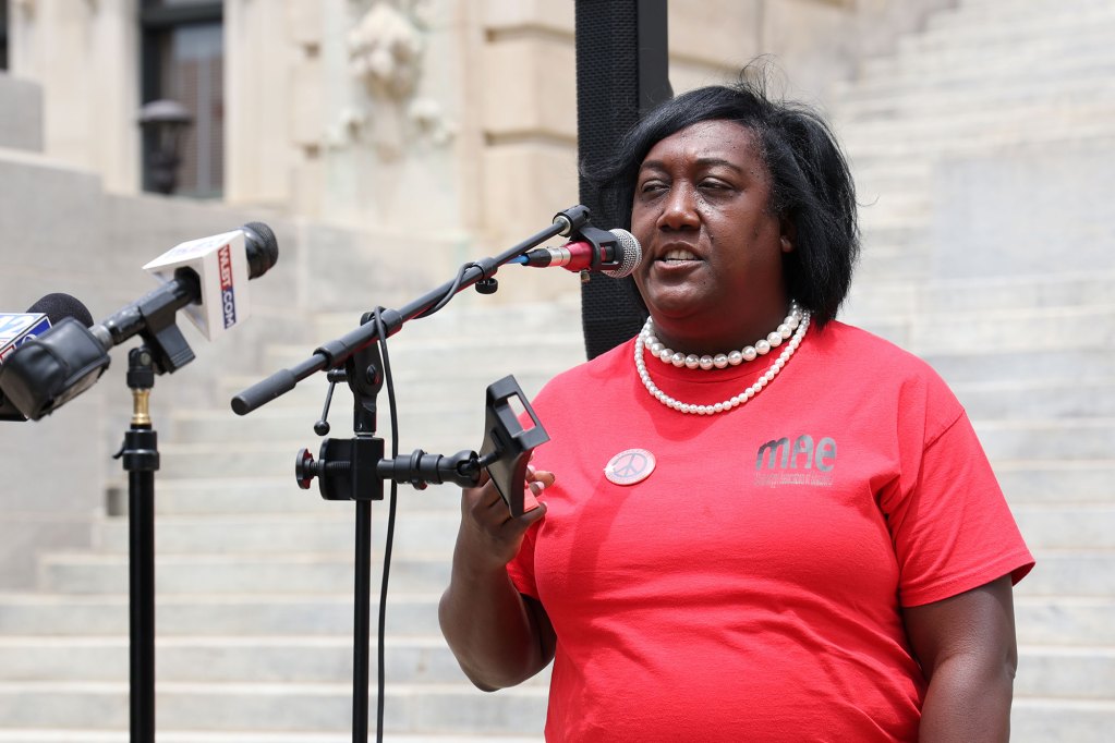 A woman in a red MAE shirt and pearl necklace speaks on an outdoor staircase